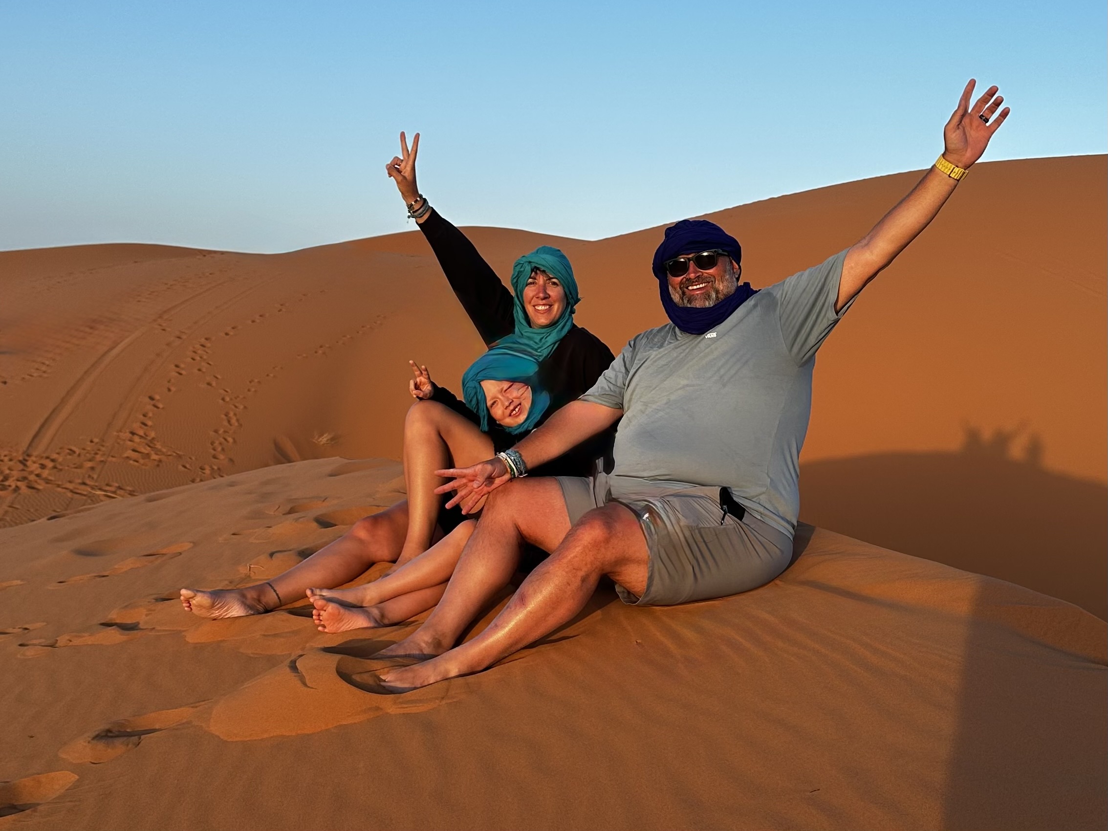 Jax Family Travels sitting on the Sahara Desert sand dunes in Morocco, arms raised